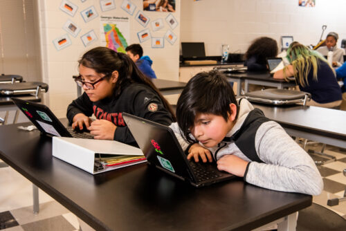 Two teenagers sit at laptops in a computing classroom.