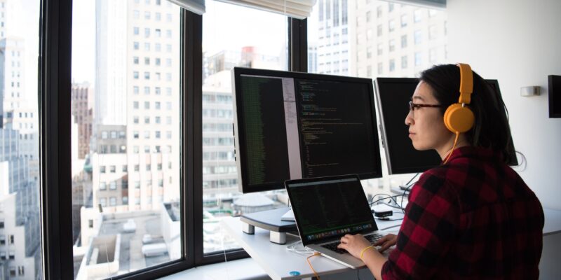 A woman works at a multi-screen computer setup on a standing desk.