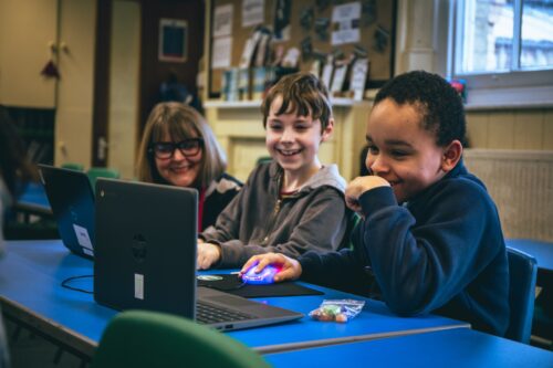 Two young people smiling whilst working on their laptop with an adult mentor by their side.