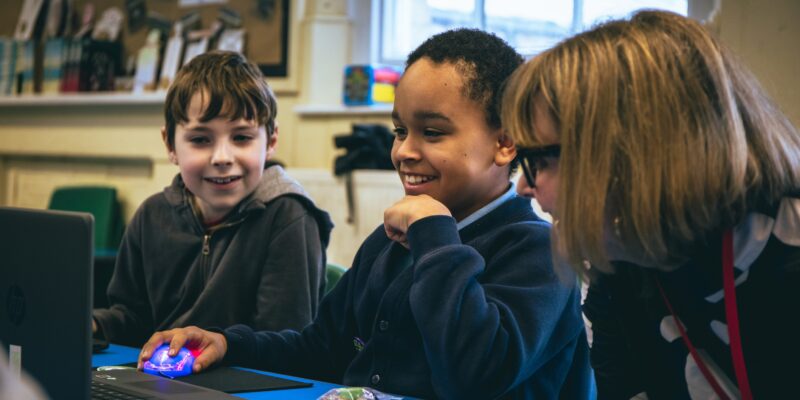 An educator helps two young learners with a coding project in a classroom.