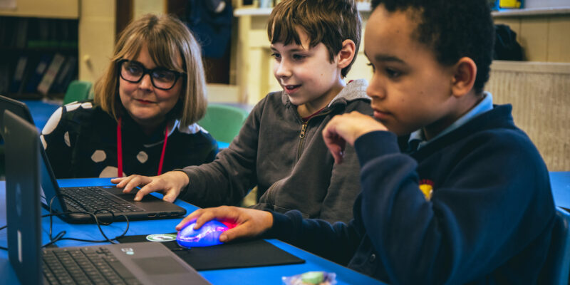 Two children code on laptops while an adult supports them.