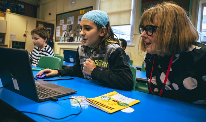 A girl codes at a laptop while a woman looks on during a Code Club session.