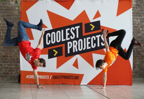 A Coolest Projects sign with two people doing handstands in front of it.