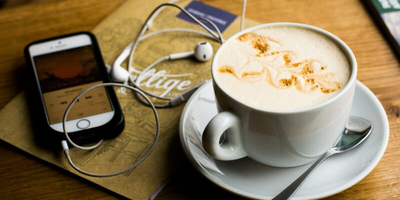 A phone with headphones plugged in next to a cup of coffee on a table.