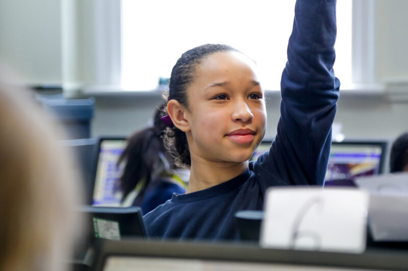 In a computing classroom, a smiling girl raises her hand.