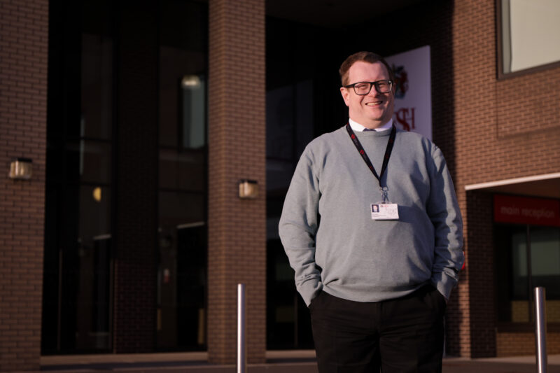 A smiling computer science teacher stands in front of a school building.