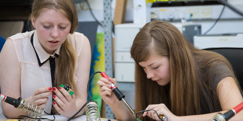 Two girls solder physical computing components in a workshop.